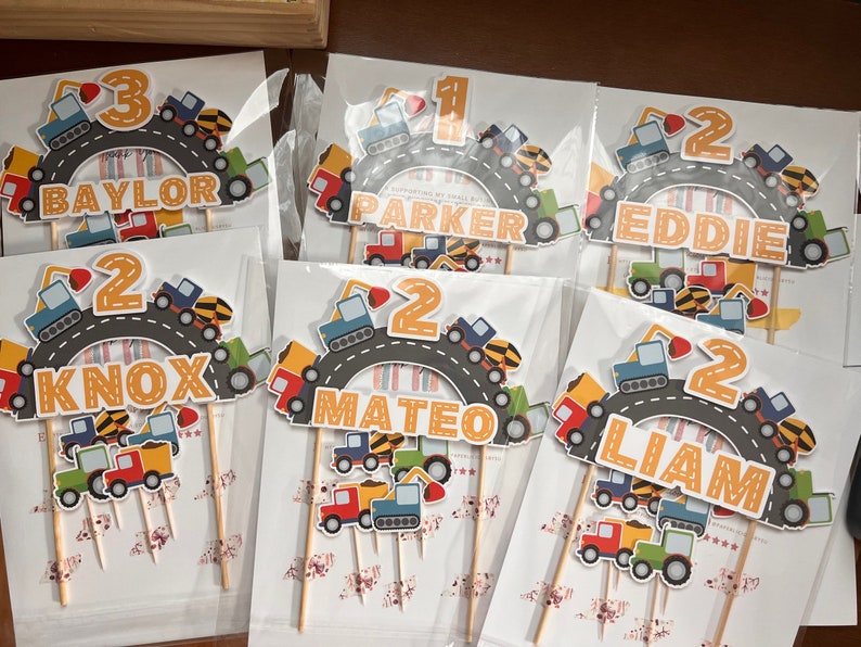 May include: Six construction-themed cake toppers with yellow, orange, and blue accents. Each topper features a number, a name, and a road with construction vehicles. The names are Baylor, Parker, Eddie, Knox, Mateo, and Liam.