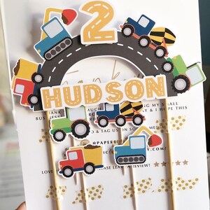 May include: Construction vehicle themed cake topper set with a yellow number 2, the name Hudson, and various construction vehicles in blue, yellow, red, and green. The vehicles are on a grey road with white lines.