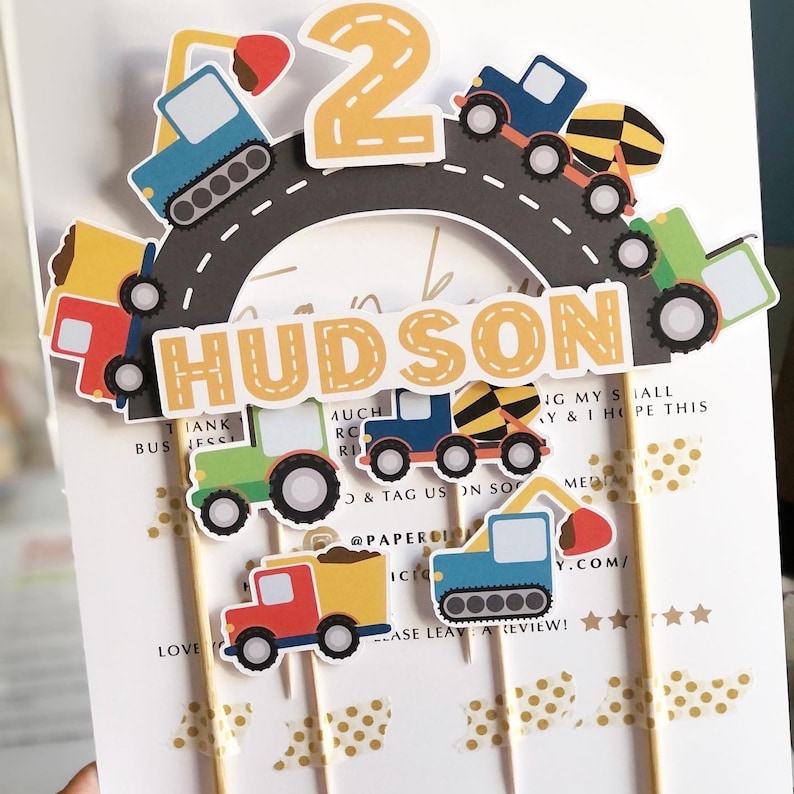May include: Construction vehicle themed cake topper set with a yellow number 2, the name Hudson, and various construction vehicles in blue, yellow, red, and green. The vehicles are on a grey road with white lines.