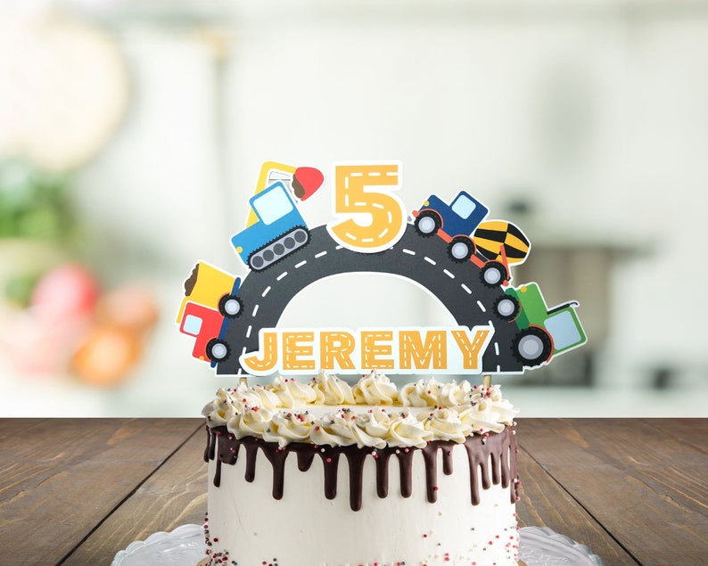 May include: A birthday cake with a construction vehicle themed cake topper. The topper features a yellow number 5, a road, and several construction vehicles. The cake is decorated with chocolate frosting and red and white sprinkles. The cake topper says "JEREMY".