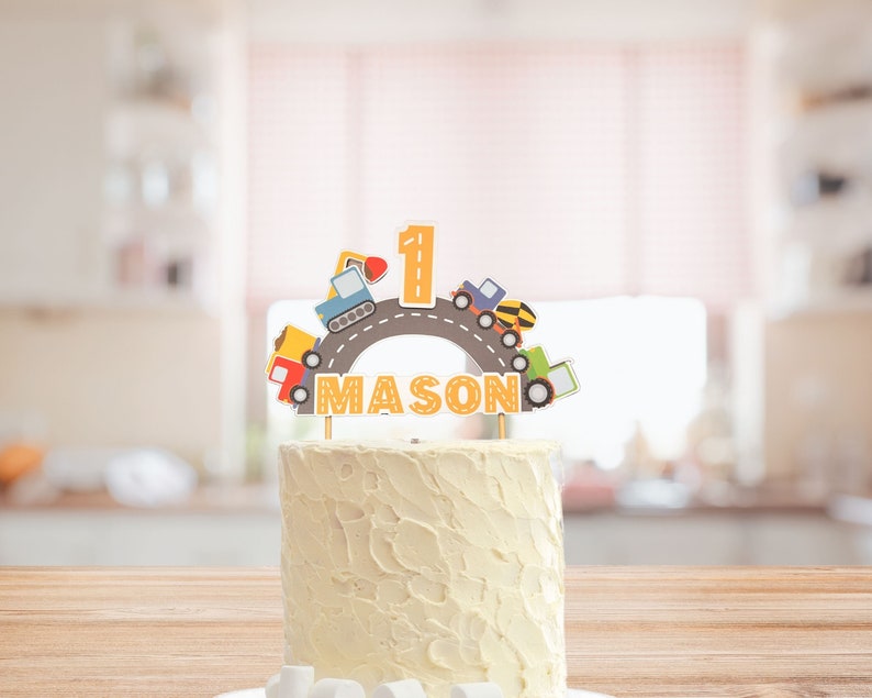 May include: A white cake with a construction-themed topper. The topper features a yellow road with a number one, a blue truck, a yellow excavator, and the name "MASON".