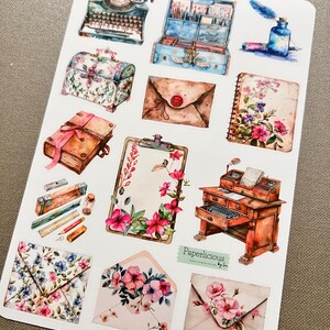 May include: A sheet of vintage-style stickers featuring images of a typewriter, a vintage suitcase, a floral-patterned box, envelopes, a book, a clipboard, a desk with a typewriter, and an inkwell with a quill pen. The stickers are printed on white paper and have a watercolor-like appearance. The text "Paperlicious by Sue" is printed in the bottom right corner.