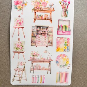 K&ouml;nnte beinhalten: Eine Bogen mit Aufklebern, die verschiedene Kunstmaterialien zeigen, darunter Pinsel, Farbtuben, eine Palette, Kreiden und eine Staffelei. Die Aufkleber sind im Aquarell-Stil mit rosa und wei&szlig;en Blumen gehalten.
