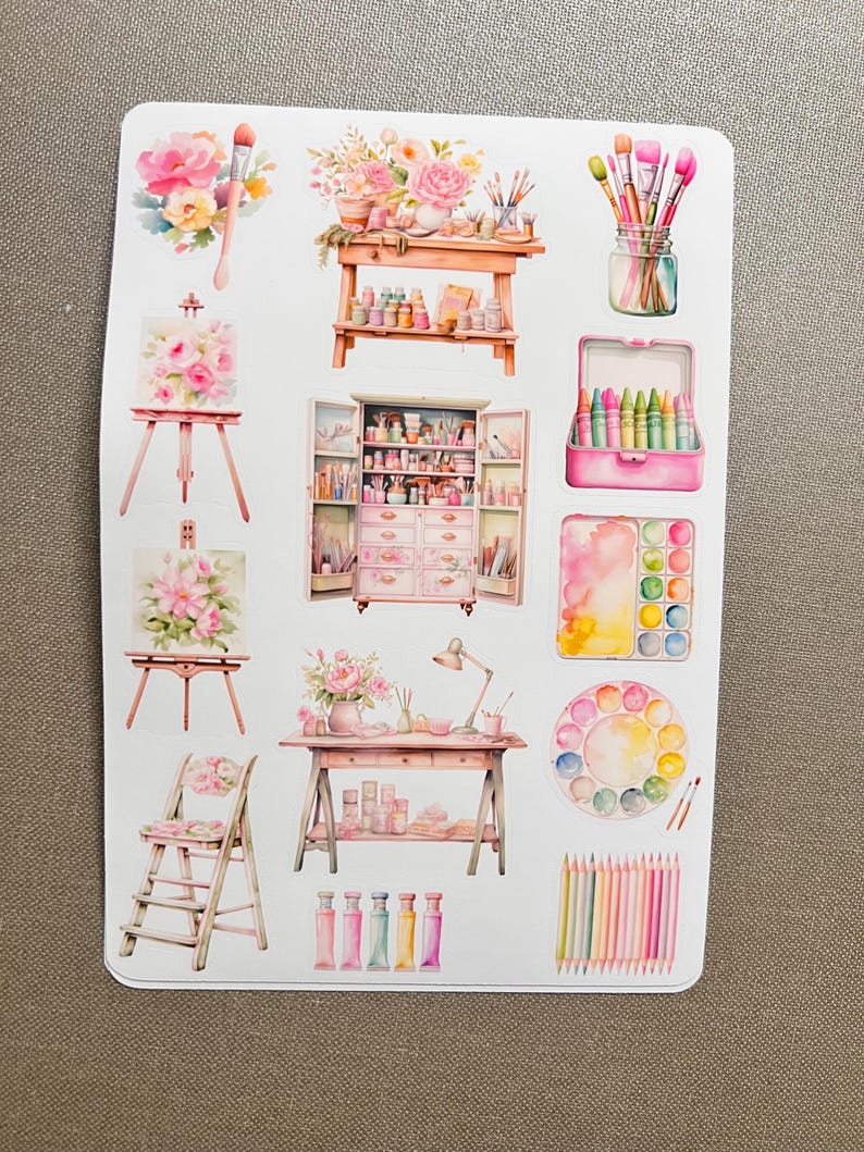 K&ouml;nnte beinhalten: Eine Bogen mit Aufklebern, die verschiedene Kunstmaterialien zeigen, darunter Pinsel, Farbtuben, eine Palette, Kreiden und eine Staffelei. Die Aufkleber sind im Aquarell-Stil mit rosa und wei&szlig;en Blumen gehalten.