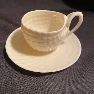 May include: Cream-colored teacup and saucer set. The cup has a textured, basketweave design and a curved handle. The saucer is round with a subtle, concentric pattern. The set is made of ceramic and is suitable for serving tea or coffee.