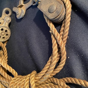 May include: An antique pulley system with a weathered appearance. The metal components show signs of age and use, with a rope threaded through the pulley. The rope is a natural tan color, knotted and looped.