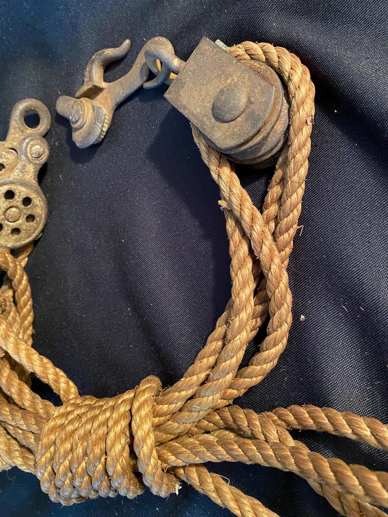 May include: An antique pulley system with a weathered appearance. The metal components show signs of age and use, with a rope threaded through the pulley. The rope is a natural tan color, knotted and looped.