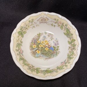 Puede incluir: Un plato pequeño, blanco, con borde festoneado y diseño floral. El plato presenta una ilustración central de flores amarillas y una pequeña figura, rodeada por un borde de hojas y flores verdes. Las palabras "Brambly Hedge Spring" están impresas en el borde.