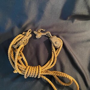May include: A vintage pulley system with a rope and metal components. The rope is a light brown color, and the metal parts have a weathered, antique appearance. The system is coiled and ready for use.