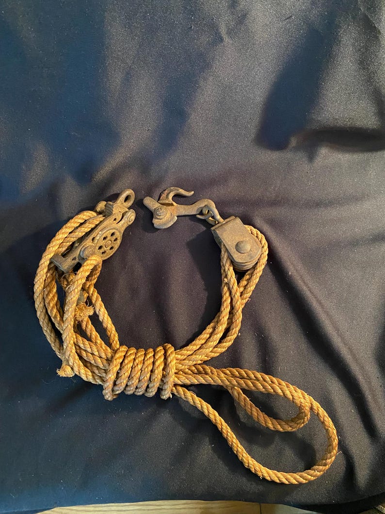 May include: A vintage pulley system with a rope and metal components. The rope is a light brown color, and the metal parts have a weathered, antique appearance. The system is coiled and ready for use.