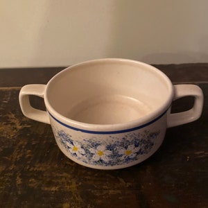 Op de afbeelding: Witte keramische soepkom met twee handvatten. De kom heeft een blauwe band rond de rand en een bloemenpatroon van blauwe en gele bloemen. De kom staat op een donker houten oppervlak.