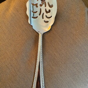 May include: A silver slotted serving spoon with a decorative handle. The spoon has a scalloped edge and a pattern of holes in the bowl.