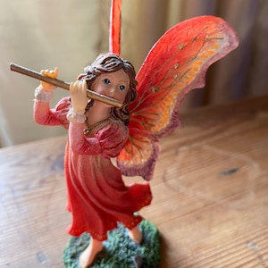 Puede incluir: Una figurita de un hada tocando una flauta. El hada tiene el pelo largo y castaño, ojos azules y lleva un vestido rojo. Tiene alas grandes, de color naranja y rosa con detalles dorados.