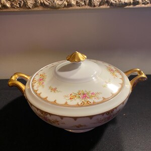Puede incluir: Un tazón de servir de porcelana blanca con tapa, con un patrón floral en tonos de rosa, amarillo y verde. El tazón tiene detalles dorados alrededor del borde y las asas.