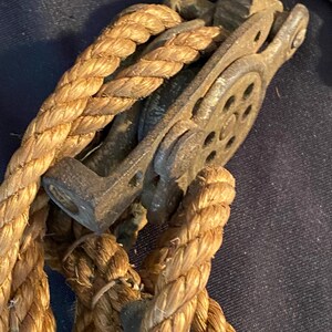 May include: Close-up of a vintage pulley system with a thick, woven rope. The pulley is made of weathered metal, showing signs of age and use. The rope is a natural tan color, tightly wound around the pulley's wheel. The image evokes a sense of nautical or industrial history.