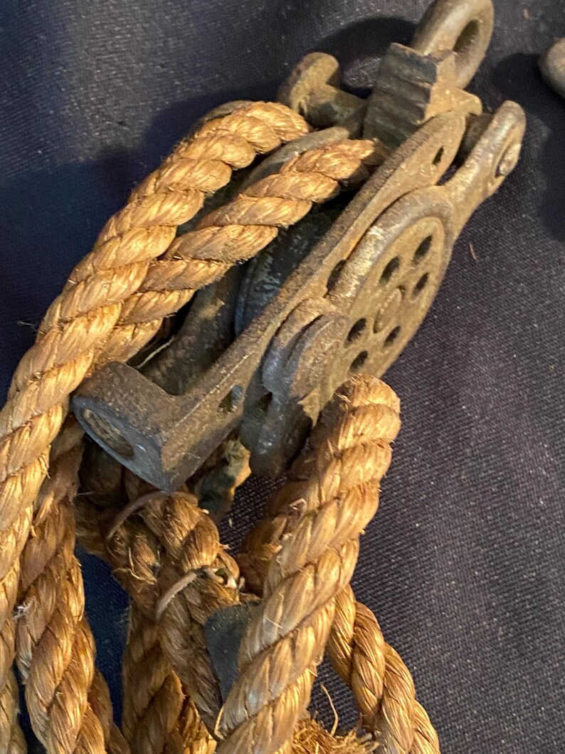 May include: Close-up of a vintage pulley system with a thick, woven rope. The pulley is made of weathered metal, showing signs of age and use. The rope is a natural tan color, tightly wound around the pulley's wheel. The image evokes a sense of nautical or industrial history.