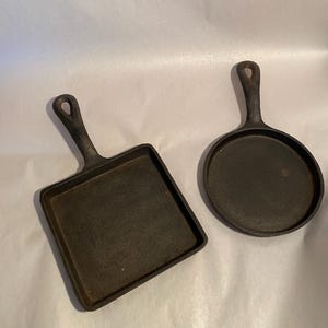 Puede incluir: Dos pequeñas sartenes de hierro fundido. Una es cuadrada, la otra redonda. Ambas tienen un asa con un agujero para colgar. Las sartenes son de color marrón oscuro y parecen bien sazonadas, listas para cocinar.