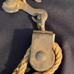 May include: Close-up of a vintage metal pulley system with a hook and rope. The pulley is made of aged, rusty metal, and the rope is thick and tan. The hook is curved and attached to the pulley.