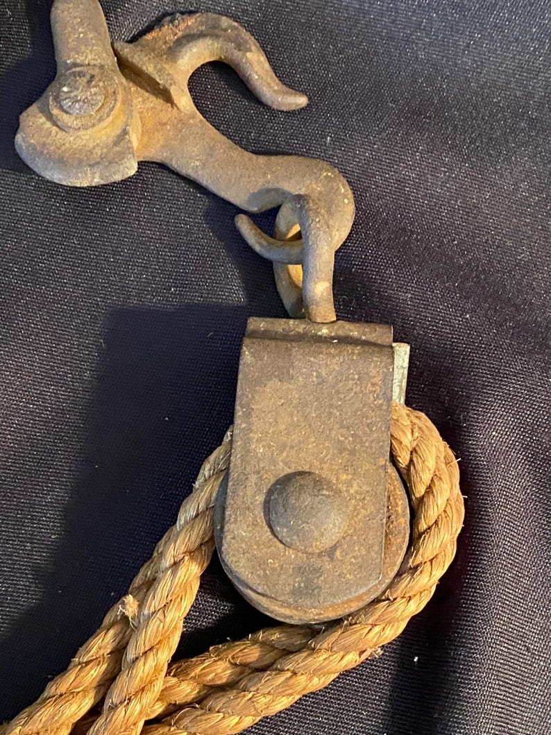 May include: Close-up of a vintage metal pulley system with a hook and rope. The pulley is made of aged, rusty metal, and the rope is thick and tan. The hook is curved and attached to the pulley.