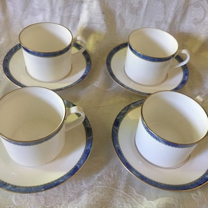 Puede incluir: Cuatro tazas y platillos blancos. Cada taza y platillo tiene un diseño de borde azul y dorado. Las tazas tienen un borde dorado y un asa pequeña. Los platillos son redondos y tienen un borde azul y dorado a juego.