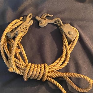 May include: An antique pulley system with a rope and metal components. The rope is tan and knotted, and the metal pieces are a tarnished bronze color. The system includes a pulley wheel and a hook.