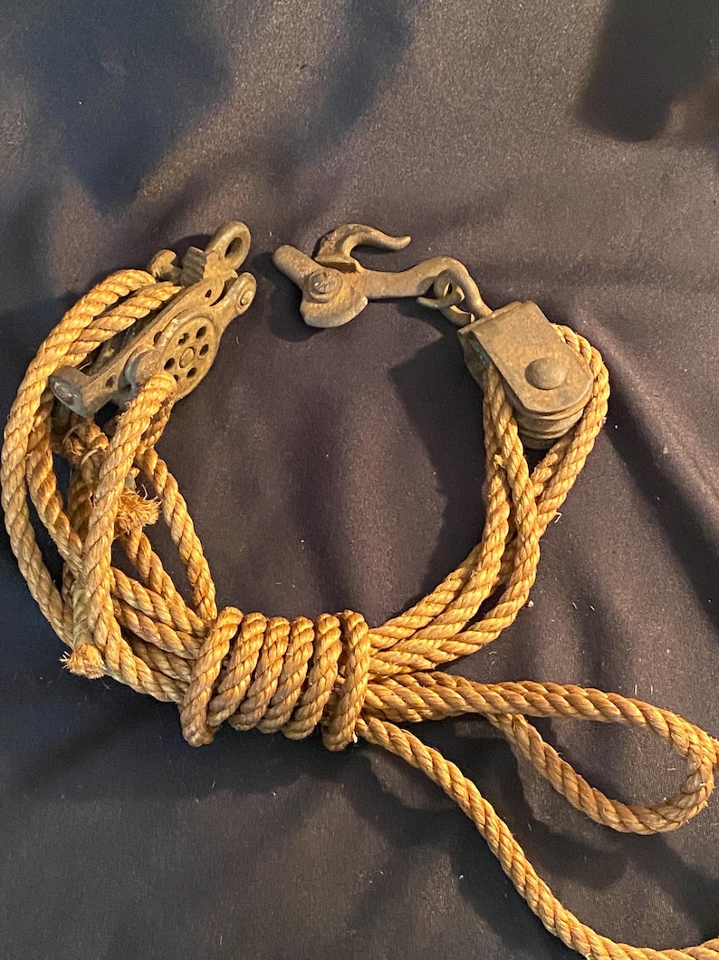 May include: An antique pulley system with a rope and metal components. The rope is tan and knotted, and the metal pieces are a tarnished bronze color. The system includes a pulley wheel and a hook.
