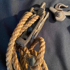 May include: Close-up of antique nautical hardware, including a weathered metal pulley and a rope. The rope is thick and twisted, with a natural tan color. The metal components show signs of age and wear, suggesting a vintage origin.