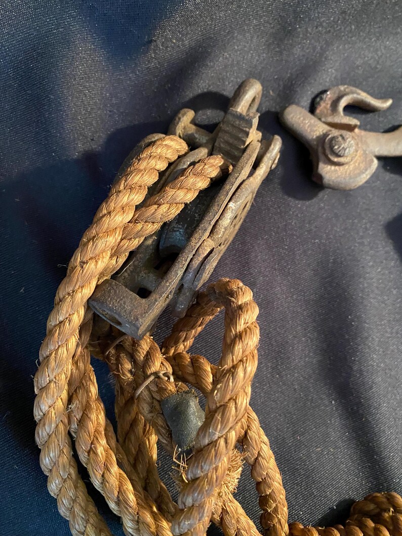 May include: Close-up of antique nautical hardware, including a weathered metal pulley and a rope. The rope is thick and twisted, with a natural tan color. The metal components show signs of age and wear, suggesting a vintage origin.