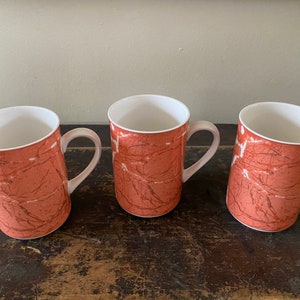 May include: Three ceramic mugs with a reddish-brown marbled design and white interiors. Each mug has a curved handle and a white interior. The mugs are arranged on a dark wooden surface.