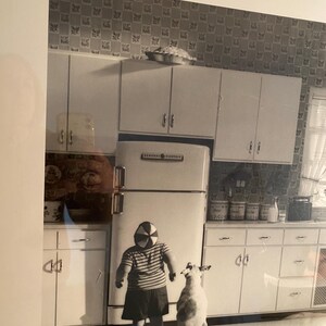 Può includere: Fotografia in bianco e nero di una scena di cucina vintage. Un bambino con una camicia a righe e una gonna è di fronte a un cane. Un frigorifero e degli armadietti riempiono lo sfondo. Una torta è sopra gli armadietti.