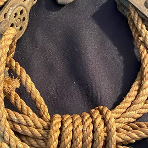 May include: Close-up of a weathered, antique pulley system with a thick, tan rope. The metal components show signs of age and rust. The rope is tightly coiled and knotted, suggesting a nautical or industrial theme.