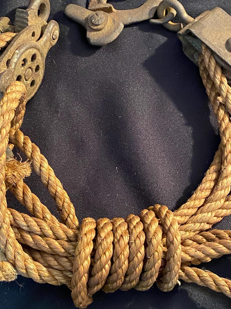 May include: Close-up of a weathered, antique pulley system with a thick, tan rope. The metal components show signs of age and rust. The rope is tightly coiled and knotted, suggesting a nautical or industrial theme.