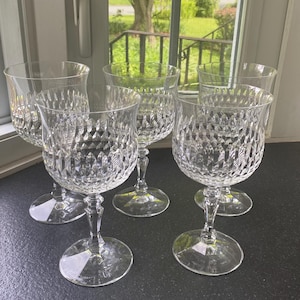 May include: Five clear crystal wine glasses with diamond-cut patterns. The glasses have a rounded bowl shape, a slender stem, and a circular base. The glasses are arranged on a dark surface near a window.