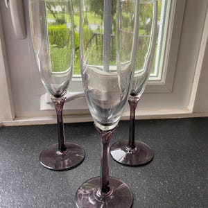 May include: Three clear glass champagne flutes with tall, slender bowls and deep purple stems and bases. Each glass features a smooth, transparent upper section that transitions into a solid, dark purple glass stem and a wide, circular base.