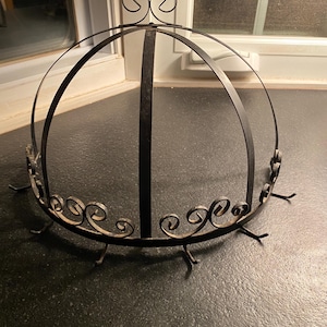 May include: A black metal decorative stand with a domed top and decorative scrollwork. The stand has a central vertical support and several small feet. The top features a decorative swirl design.