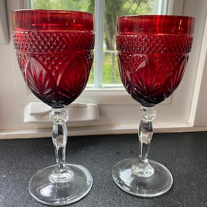 May include: Two matching wine glasses featuring deep red bowls with a textured diamond pattern on the upper section and a pressed leaf design on the lower section. Each glass has a clear, faceted stem and a round, transparent glass base.