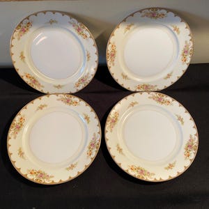 Puede incluir: Cuatro platos de porcelana blanca con diseño floral y ribete dorado. Los platos tienen un borde festoneado y un delicado patrón de flores rosas, amarillas y verdes. Los platos están colocados sobre una superficie negra.