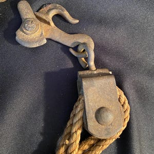 May include: Close-up of antique metal pulley system with a hook and rope. The metal components show signs of age and wear, with a rusty brown patina. The rope is a thick, twisted, natural fiber in a light brown color.