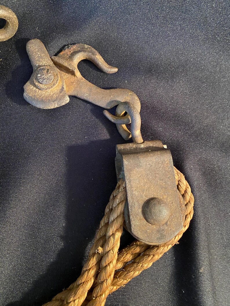 May include: Close-up of antique metal pulley system with a hook and rope. The metal components show signs of age and wear, with a rusty brown patina. The rope is a thick, twisted, natural fiber in a light brown color.