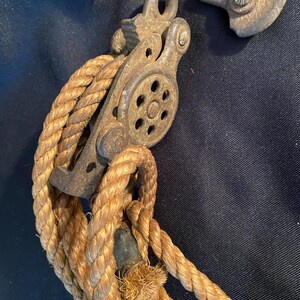 May include: Close-up of a vintage pulley system with a thick, tan rope. The pulley is made of weathered metal, showing signs of age and use. The rope is tightly wound around the pulley wheel, suggesting a mechanism for lifting or pulling heavy objects. The background is a dark blue fabric.