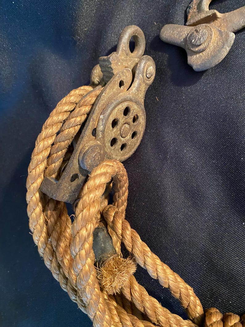 May include: Close-up of a vintage pulley system with a thick, tan rope. The pulley is made of weathered metal, showing signs of age and use. The rope is tightly wound around the pulley wheel, suggesting a mechanism for lifting or pulling heavy objects. The background is a dark blue fabric.