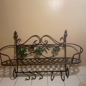 Op de afbeelding: Oornamentale metalen wandplank met een bronzen afwerking. Voorzien van een plank met een traliewerkontwerp, decoratief krulwerk en druivenbladaccenten. Inclusief haken om items op te hangen. De plank is ongeveer 61 cm breed.
