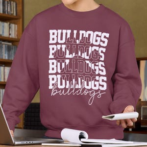 May include: A maroon sweatshirt with the word "BULLDOGS" printed in white, stacked vertically four times. The word "bulldogs" is also written in cursive at the bottom. The sweatshirt is a crew neck style.