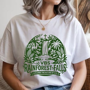 May include: White t-shirt with a green rainforest-themed graphic. The design includes a waterfall and tropical foliage within a semi-circular frame. The text reads "VBS Rainforest Falls" with the tagline "Explores the Nature of God".