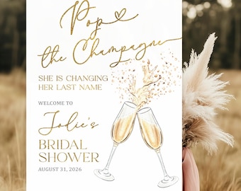 Puede incluir: Un letrero blanco sobre un caballete de madera con texto dorado que dice "Pop the Champagne" y "Bridal Shower de Jolie, 31 de agosto de 2026". El letrero presenta copas de champán y está decorado con hierba de la pampa y tela rosa.