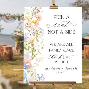 May include: A white wedding sign on a wooden easel with floral accents. The sign reads "Pick a seat, not a side. We are all family once the knot is tied." Names and a date are also included.