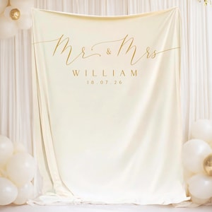 Op de afbeelding: Een crèmekleurige bruiloft achtergrond met gouden tekst "Mr. & Mrs. WILLIAM 18.07.26". De achtergrond is gedrapeerd en omgeven door witte en gouden ballonnen, wat een feestelijke sfeer creëert voor een bruiloft of speciaal evenement.