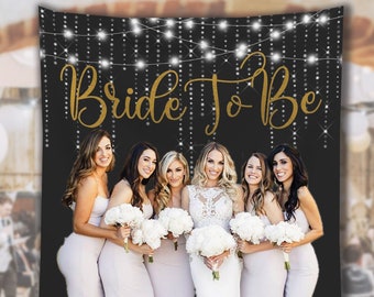 May include: A black backdrop with gold text that reads "Bride To Be" and string lights. Six women in light-colored dresses hold bouquets of white flowers. The backdrop size is 88"x104".