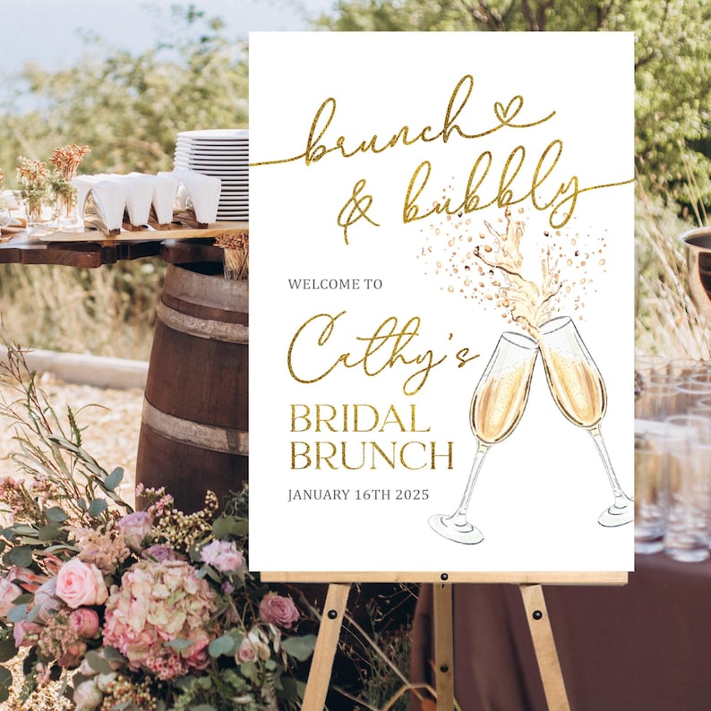 May include: A white sign on a wooden easel with gold lettering that reads "brunch & bubbly". Below, it says "Welcome to Cathy's Bridal Brunch January 16th 2025". Two champagne glasses are illustrated.
