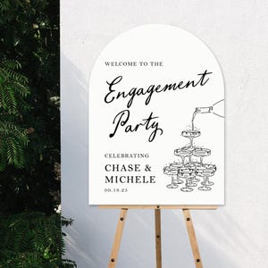 K&ouml;nnte beinhalten: Ein wei&szlig;es Schild mit schwarzem Text, das eine Verlobungsfeier f&uuml;r Chase & Michelle am 18. September 2025 ank&uuml;ndigt. Das Schild zeigt eine Grafik einer Champagnerpyramide mit einer Flasche, die Champagner in das oberste Glas gie&szlig;t.